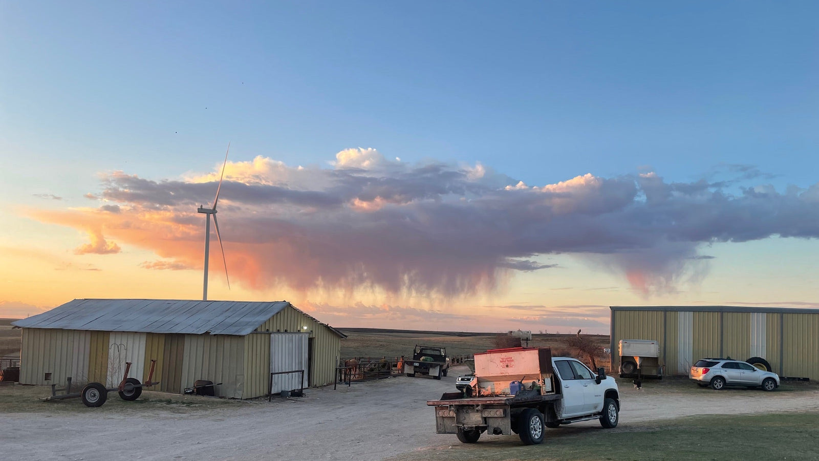Truck with a trailer in a rural setting with a windmill and buildings under a colorful sky.